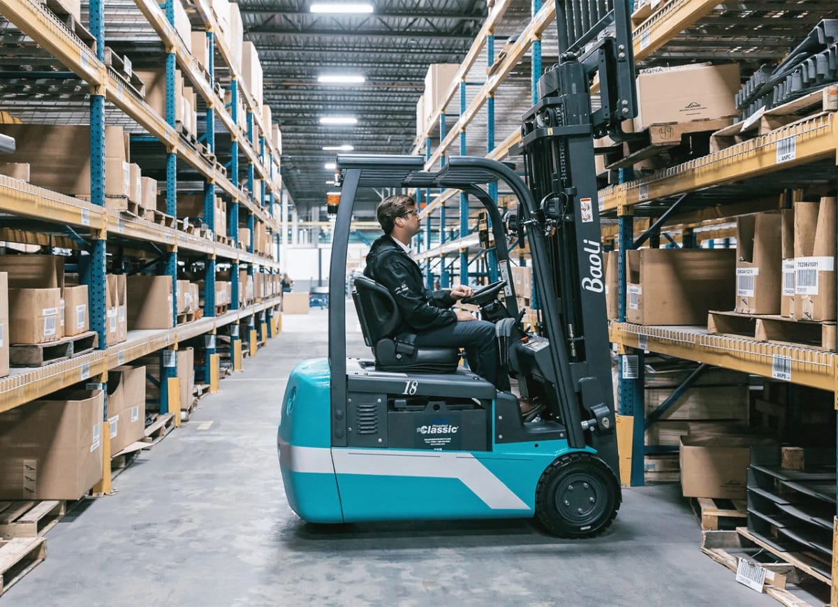Baoli_electric_forklift_truck_kbe_t15-18-20_content-jpg | Jović Viljuškari Radnik upravlja tirkiznim viljuškarom niz prolaz skladišta obložen visokim paletama kutija na metalnim policama