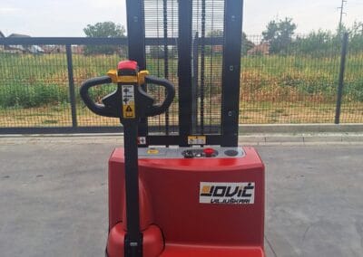 staxx-ws15h-3 | Jović Viljuškari Red electric pallet stacker with a tall black mast and yellow control handle, on a concrete surface outdoors near a fence.