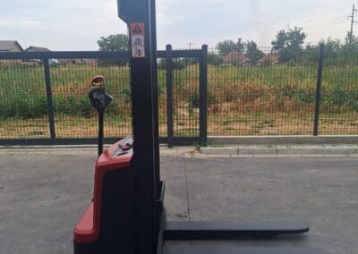 staxx-ws15h-4 | Jović Viljuškari Electric pallet stacker with red base and long black mast, forks extended forward, positioned on a concrete surface outdoors; signs are visible on the mast beside a fence and open field in the background.